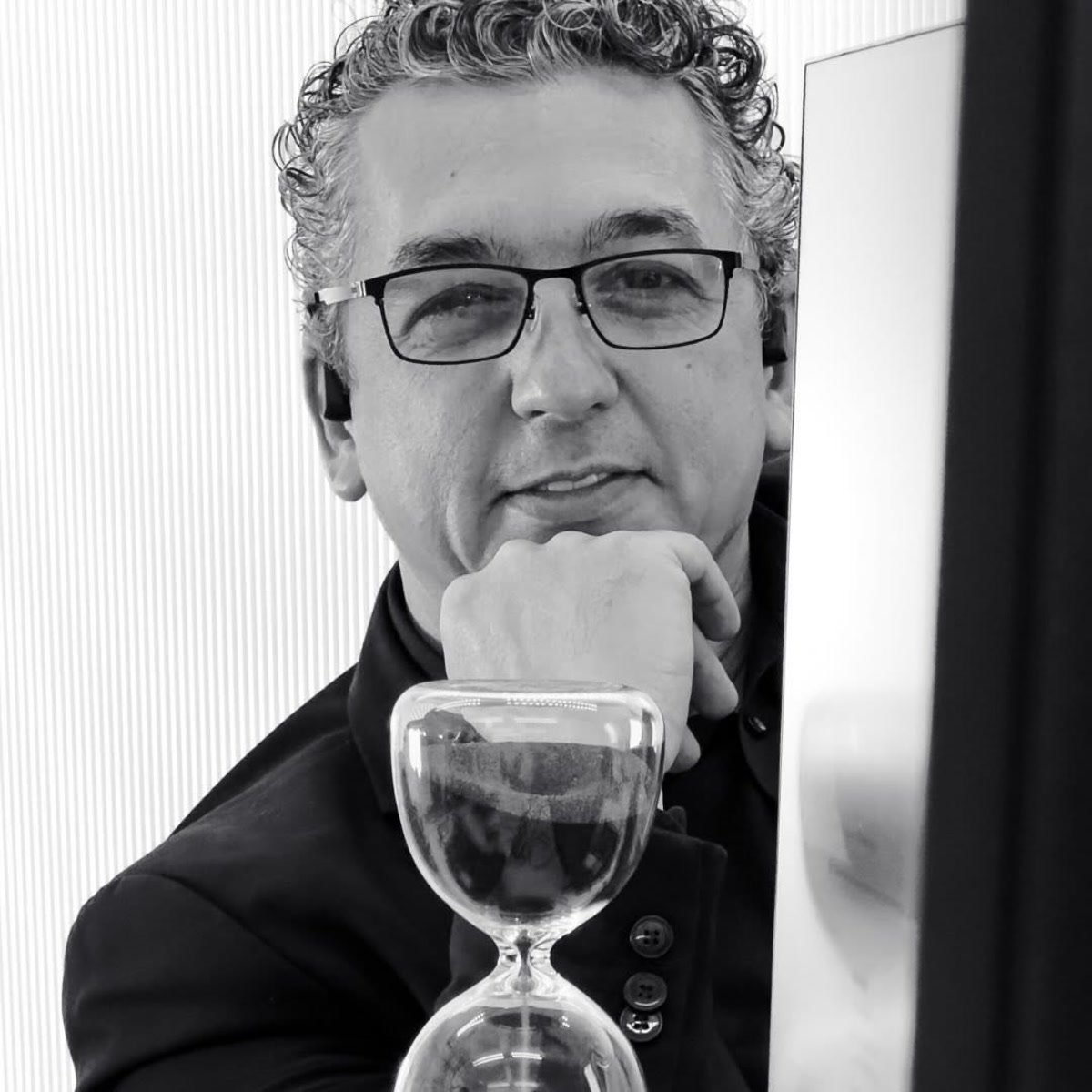 George Sebastian, black-framed glasses, black shirt, chin resting on his fist beside an hourglass